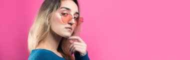 Son moda kalp gözlüklü, utangaç bir gülümsemeyle poz veren romantik sarışın kız. Pembe arka planda stüdyo portresi.