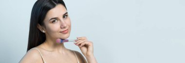 Açık renk bluzlu, beyaz arka planda yüz makyajı yapan genç bir kızın yakın çekimi. Güzel bir kadın yüzüne bir makyaj fırçası tutar ve gülümser..