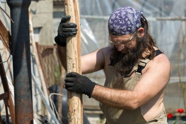 Bandanalı orta yaşlı sakallı bir adam baltayla kütük kesiyor. Tulumlu gaddar sıcak bir günde zor işi yapar..