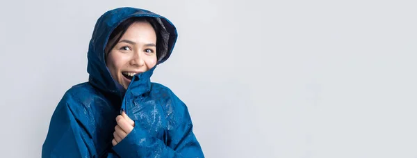 Portrait of a smiling girl dressed in blue raincoat in drops posing with hood on grey background in a studio.