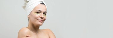 Kafasında beyaz havluyla gezen genç bir kız gözlerinin altına kolajen jel bantlar takıyor. Göz teması altında maske.