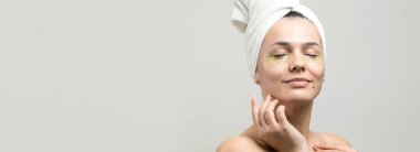 Kafasında beyaz havluyla gezen genç bir kız gözlerinin altına kolajen jel bantlar takıyor. Göz teması altında maske.