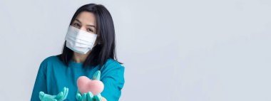 Koruyucu tıbbi maske takan esmer genç bir kız pembe bir kalp taşıyor, hayatı simgeliyor. Coronavirus konsepti.