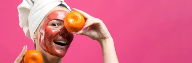 Kafasında kırmızı besleyici maske olan beyaz havluyla güzel bir kadın portresi. Cilt temizleme organik kozmetik rahatlama konsepti. Sırtı turuncu bir mandalina tutan bir kız..
