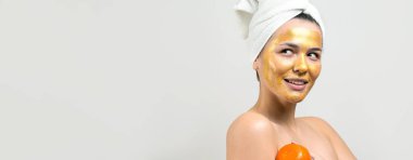 Yüzünde altın besleyici maske olan beyaz havluyla kadın portresi. Cilt temizleme organik kozmetik rahatlama konsepti. Sırtı turuncu bir mandalina tutan bir kız..