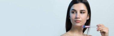 Açık renk bluzlu, beyaz arka planda yüz makyajı yapan genç bir kızın yakın çekimi. Güzel bir kadın yüzüne bir makyaj fırçası tutar ve gülümser..