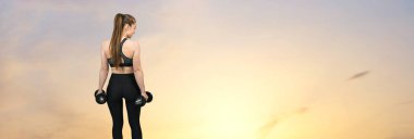 Spor giyimli genç ve zayıf bir atletik kız deniz kenarında gün batımına karşı bir takım egzersizler yapıyor. Dambıllarla egzersiz yapar. Fitness ve sağlıklı yaşam tarzı.