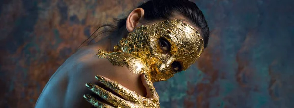 Girl with a mask on her face made of gold leaf. Gloomy studio portrait of a brunette on an abstract background.