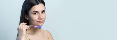 Açık renk bluzlu, beyaz arka planda yüz makyajı yapan genç bir kızın yakın çekimi. Güzel bir kadın yüzüne bir makyaj fırçası tutar ve gülümser..