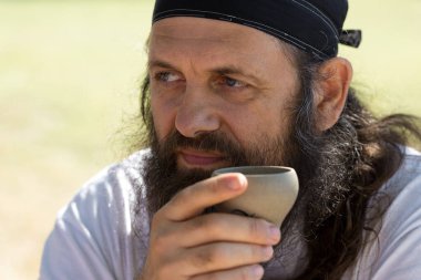 Bandanalı sakallı bir adam doğanın arka planında çay içiyor..