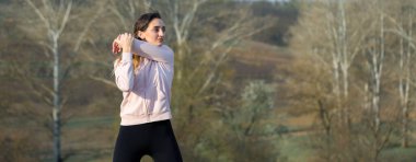 Spor giyimli genç, zayıf, atletik bir kız bir takım egzersizler yapıyor. Yeşil bahar otlak tepelerinin arka planında sağlık ve sağlıklı yaşam tarzı.