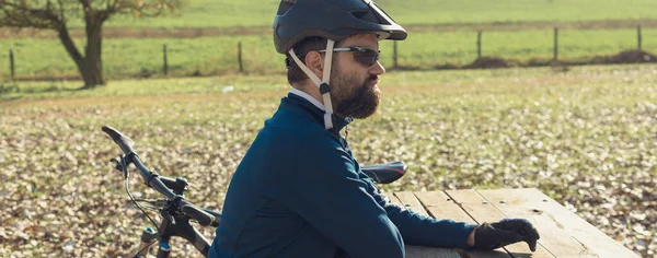 Cyclist in pants and fleece jacket on a modern carbon hardtail bike with an air suspension fork rides off-road. The guy is resting on a bench in the autumn park.