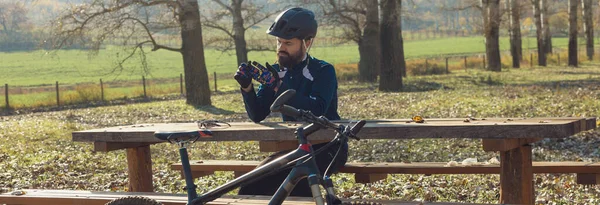 Cyclist in pants and fleece jacket on a modern carbon hardtail bike with an air suspension fork rides off-road. The guy is resting on a bench in the autumn park.