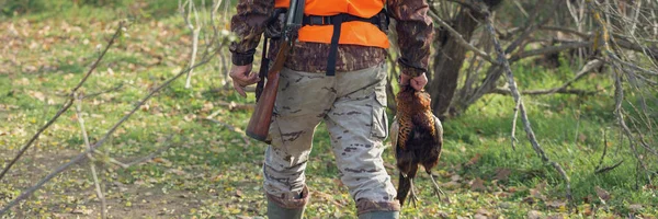 手に銃を持った男と曇りの日に森の中でキジ狩りをするオレンジのベスト 彼の手にキジがあるハンター — ストック写真