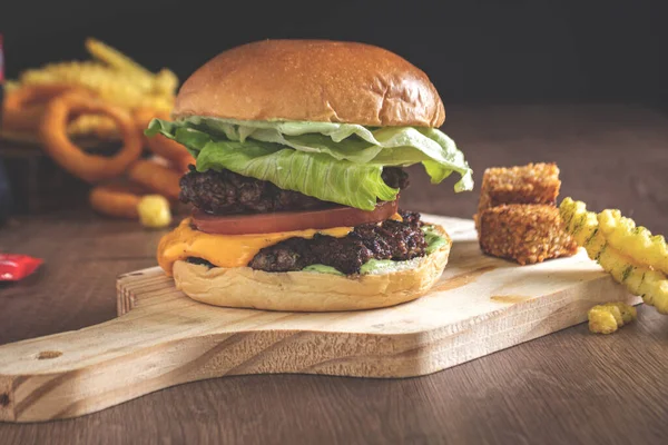 double cheeseburger with bacon lettuce, tomato, onion in hamburger buns on a wooden board on a black background with french fries in the background