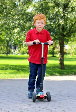 Çocuk güneşli yaz gününde bir şehir parkında scooter binmek öğrenirler. Bir scooter ile poz zencefil saçlı neşeli küçük çocuk. Çocuklar için aktif eğlence ve açık hava sporu