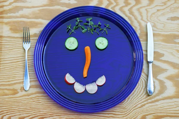 A blue glass plate with a vegetable face. Cucumbers, carrots, radishes and oregano on a plate served as a face.