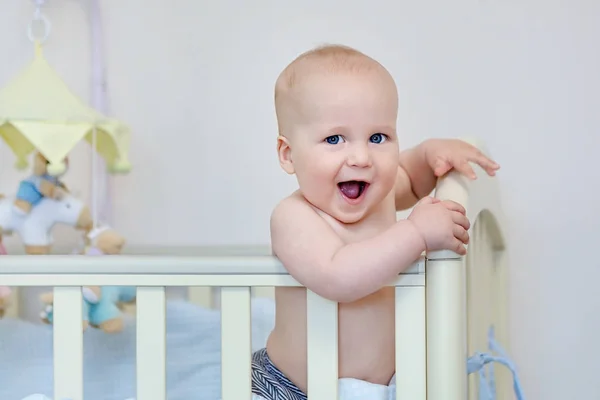 Little funny boy standing in baby bed and laughing. Smiling adorable child on feet in playpen. Kid portrait at nursery. Happy childhood concept — Stock Photo, Image