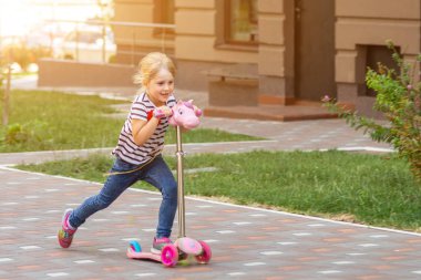 Sevimli küçük kız sürme scooter şehir park parlak yaz gününde. Sarışın yürümeye başlayan çocuk sokakta eğleniyor. Etkin boş ve açık spor çocuk kavramı için