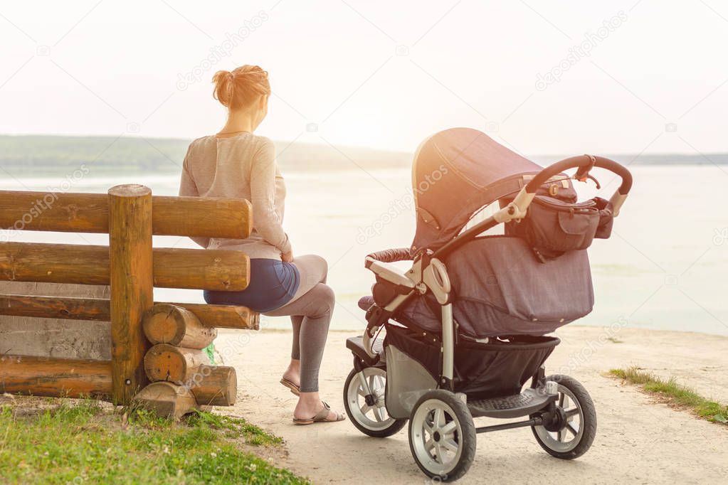 Young sporty mother with stroller sitting on wooden bench near lake or river. Mom walking with baby in pram near pond at early morning. Healthy outdoor sleeping.Toned.