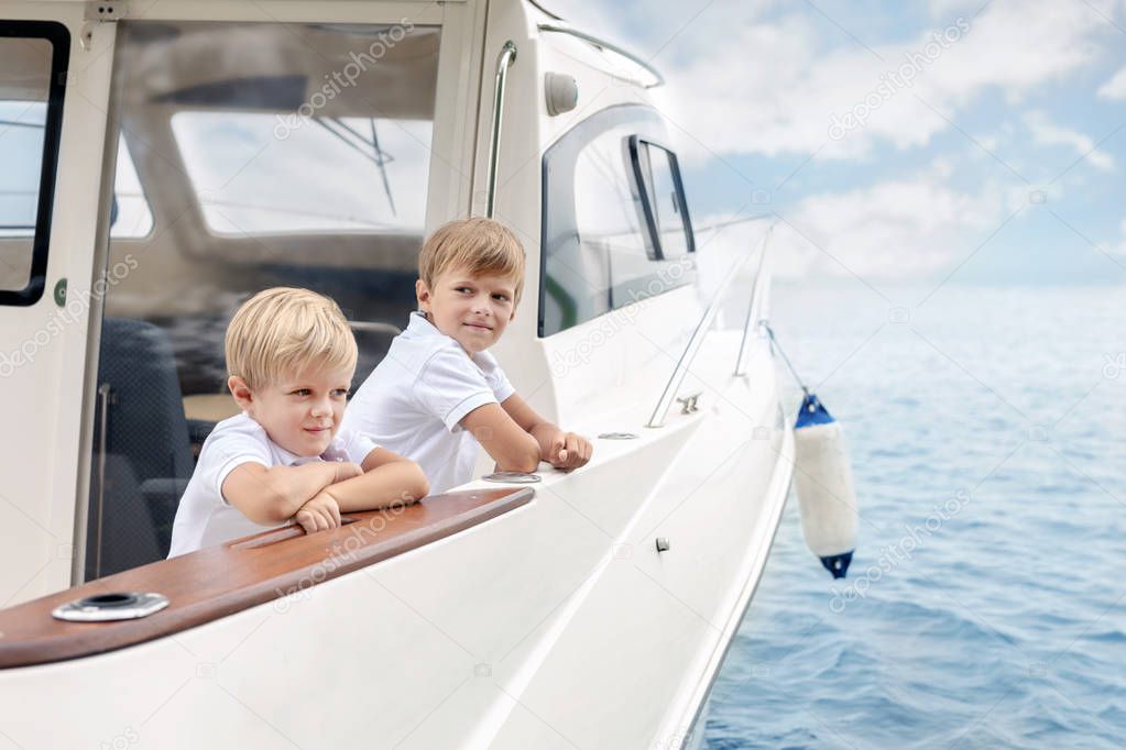 Two cute caucasian blond boys on board of white luxury yacht on bright summer day. Siblings having fun learning yachting tohether. Travel and adventure with children concept