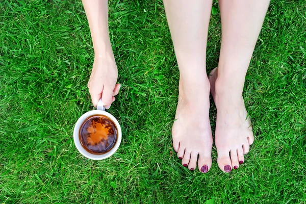 Вид сверху на ноги и руку молодой женщины, держащей чашку чая, сидящей на зеленой траве. Здравоохранение. Отдых на открытом воздухе — стоковое фото