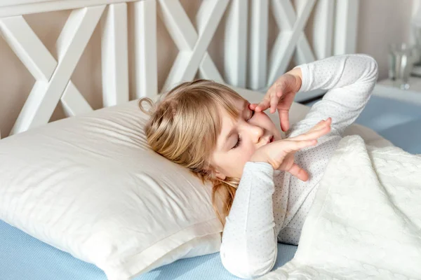 Cute little blond caucasian girl awakening at bed in morning. Child wake up early to go to school. Stretching and yawning. Healthy sleeping. Children healthcare — Stock Photo, Image