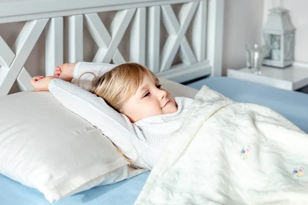 Cute little blond caucasian girl awakening at bed in morning. Child wake up early to go to school. Stretching and yawning. Healthy sleeping. Children healthcare — Stock Photo, Image