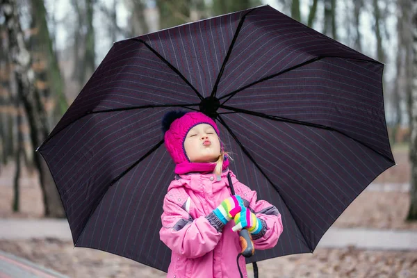 ピンクの暖かいジャケットと寒い日に都市公園における傘の下で歩いているニットの帽子を身に着けている愛らしい白人少女 — ストック写真