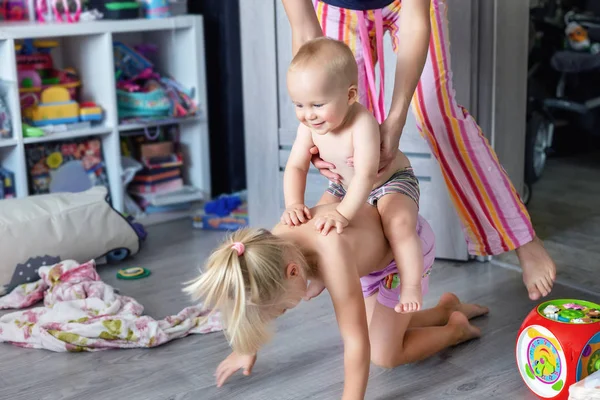 Brother and sister with mother laughing and having fun indoor.Infant boy playing, riding elder sister back like on horse. Siblings play together at playroom. Happy and careless childhood — Stock Image Brother and sister with mother laughing and having fun indoor.Infant boy playing, riding elder sister back like on horse. Siblings play together at playroom. Happy and careless childhood Royalty Free Stock Images