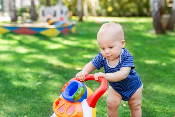 Lindo niño aprendiendo a caminar con un juguete caminante en el césped de hierba verde en el patio trasero. Bebé riendo y divirtiéndose dando el primer paso en el parque en el día soleado brillante al aire libre. Concepto de infancia feliz —  Fotos de Stock