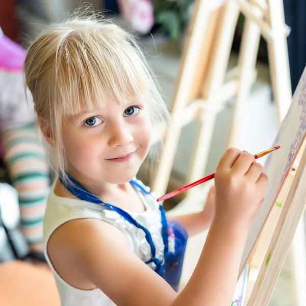 可爱的金发微笑的女孩绘画在画架在艺术工作室的讲习班课 手持刷子的孩子 用颜料画画很有趣 儿童发展概念 — 图库照片#