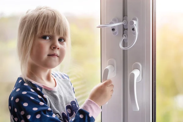 Little cute toddler girl trying to open window in apartment at high-tower building. Children window protection lock. Cable safety guard prevent opening window by child. Prevention of falling accident — Stock Photo, Image