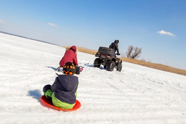 Homem em quadbike ATV andar trenós com crianças a reboque na superfície do lago congelado no inverno. Esportes radicais de inverno e recreação. Crianças ao ar livre diversão e atividades — Fotografia de Stock