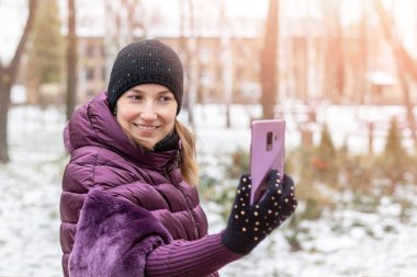 Kışlık şehir parkında yürürken akıllı telefonuyla selfie çekerken mor ceketli mutlu genç bir kadın gülümsüyor. Güzel kız kış tatillerini ve seyahatleri sever..