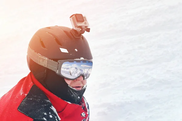 Snowboarder or skier portrait in sport goggles and protection helmet with mounted action camera and ski slope on background. Exreme winter sport outdoor activities.
