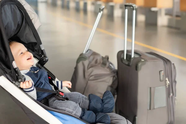 Cute funny caucasian baby boy sitting in stroller near luggage at airport terminal. Child sin carriage with suitcasese near check-in desk counter. Travelling with small children concept
