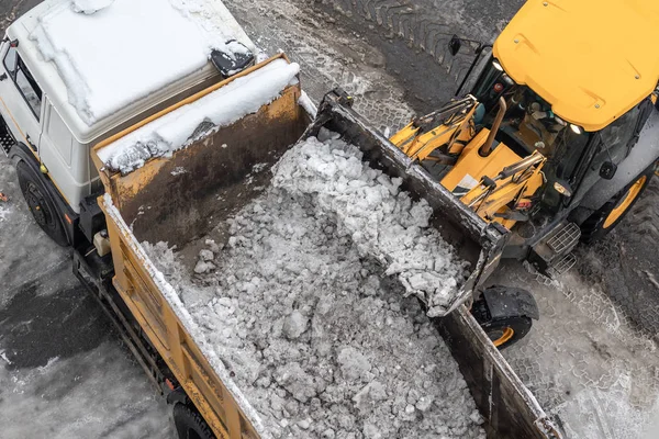 Tractor loader machine uploading dirty snow into dump truck. Cleaning city street, removing snow and ice after heavy snowfalls and blizzard