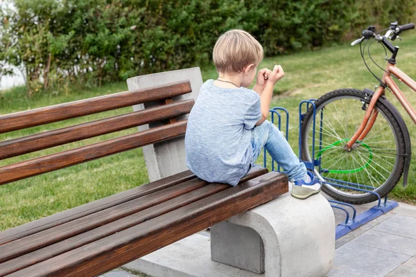 Egyedül a kis szőke ideges fiú alkalmi farmert hord, ül a fa pad a parkban. Magányos boldogtalan gyerek kint — Stock Fotó