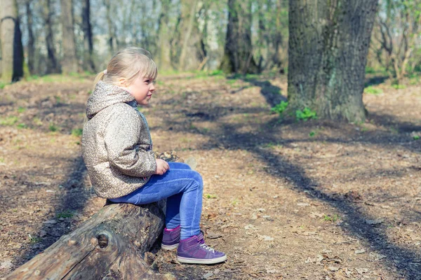 Linda chica rubia caucásica sittng en madera de tronco en el bosque y buscando en algún lugar. Adorable niño pensativo soñando con algo durante el paseo por el parque o el bosque — Foto de Stock