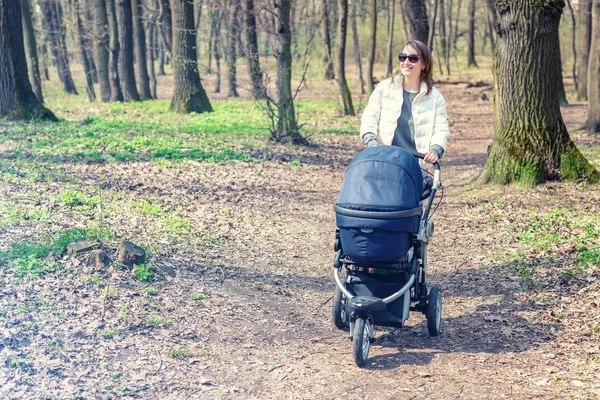 Gyönyörű fiatal felnőtt nő séta a baba babakocsi, erdőben vagy parkban fényes napsütéses nap. Egészséges életmód és a gyermekek egészségügyi ellátás. Boldog gyermekkor és a szülői — Stock Fotó