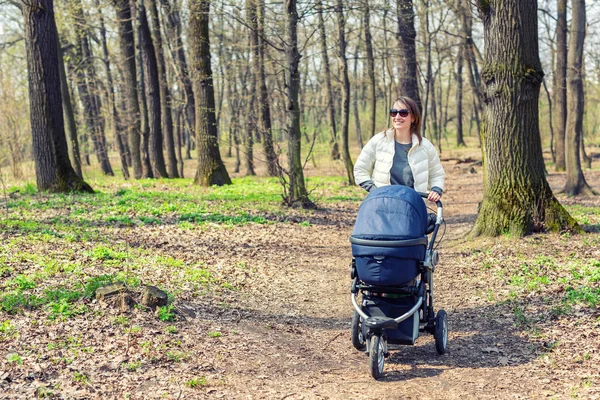 Gyönyörű fiatal felnőtt nő séta a baba babakocsi, erdőben vagy parkban fényes napsütéses nap. Egészséges életmód és a gyermekek egészségügyi ellátás. Boldog gyermekkor és a szülői — Stock Fotó