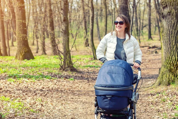 Gyönyörű fiatal felnőtt nő séta a baba babakocsi, erdőben vagy parkban fényes napsütéses nap. Egészséges életmód és a gyermekek egészségügyi ellátás. Boldog gyermekkor és a szülői — Stock Fotó