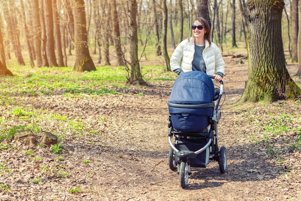 Gyönyörű fiatal felnőtt nő séta a baba babakocsi, erdőben vagy parkban fényes napsütéses nap. Egészséges életmód és a gyermekek egészségügyi ellátás. Boldog gyermekkor és a szülői — Stock Fotó