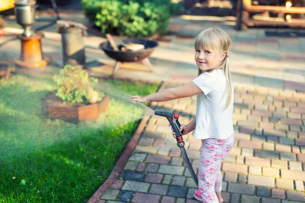 Little cute baby girl podlewanie świeże zielone trawnik mear dom podwórku na jasny letni dzień. Dziecko bawią się z tryskaczami węża wodnego — Zdjęcie stockowe