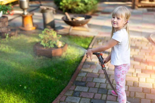 Little cute baby girl podlewanie świeże zielone trawnik mear dom podwórku na jasny letni dzień. Dziecko bawią się z tryskaczami węża wodnego — Zdjęcie stockowe