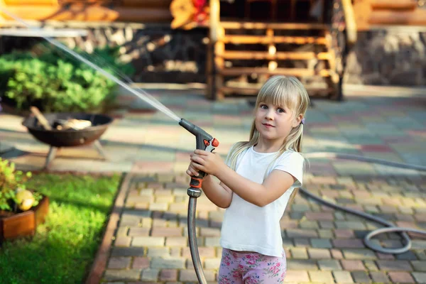 Little cute baby girl podlewanie świeże zielone trawnik mear dom podwórku na jasny letni dzień. Dziecko bawią się z tryskaczami węża wodnego — Zdjęcie stockowe