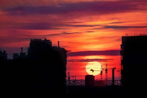 建物や建設現場のクレーンのシルエットと、夕暮れ時の大きな血まみれの赤い太陽を背景に。風光明媚な産業景観 — ストック写真
