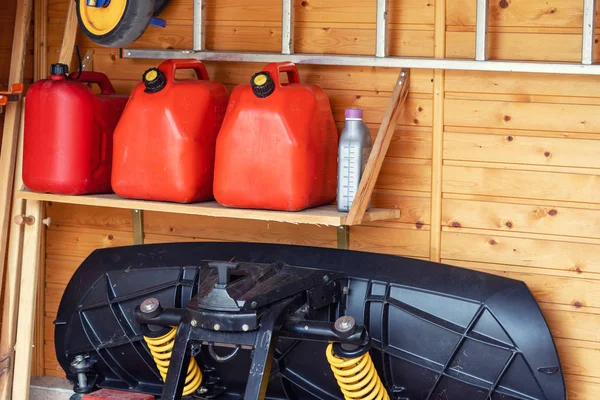 Angolo garage con tre taniche di carburante in plastica rossa, scala e spazzaneve per ATV con parete in legno su sfondo. Contenitori di gas di benzina riserve di stoccaggio a casa del veicolo garage — Foto Stock