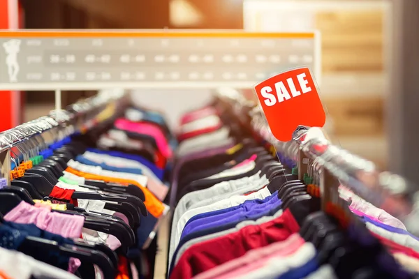 Retail clothes store clearance. Garment shop with various bright youth casual wear at discount price. Wear hangers and red SALE signboard. Seasonal sales on fashion market